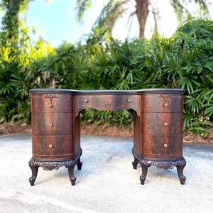 May include: A dark brown antique vanity with a curved design. The vanity has three drawers on each side, ornate detailing, and decorative hardware. The legs are curved and feature carved accents. The background includes lush green foliage and a bright sky.