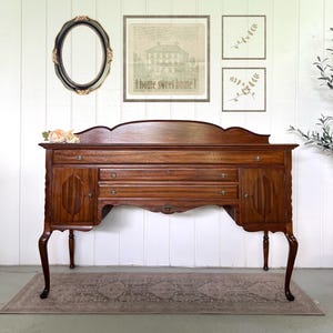 May include: A brown wooden sideboard featuring a curved backboard, two side cabinets with vertical paneling, and two central drawers. The furniture piece stands on four cabriole legs and includes small metal hardware pulls on each drawer and door.