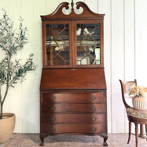 May include: A tall brown wooden secretary desk featuring a top hutch with two glass doors and a lattice grid pattern. The base includes a drop-down writing surface and four stacked drawers with curved fronts, brass hardware, and carved feet.