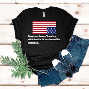 May include: Black t-shirt with a reversed American flag graphic and the text "Fascism doesn't arrive with tanks. It arrives with excuses."