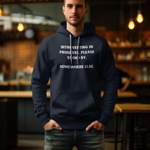May include: Navy blue hooded sweatshirt with white text that reads: "Introverting in progress. Please stand by. Somewhere else." The sweatshirt has a front pocket and a drawstring hood.