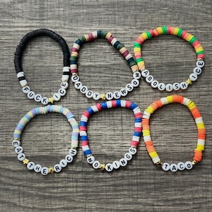 May include: Six colorful beaded bracelets with white letter beads spelling out phrases like "FUCK ICE" and "ABOLISH". The bracelets are made of flat, round beads in various colors, including black, rainbow, and orange. Each bracelet has a gold star charm.