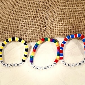 May include: Three colorful beaded bracelets with white letter beads spelling out names. One bracelet has black, yellow, and white beads, another has rainbow beads, and the third has red, white, and blue beads. The bracelets are on a white surface with a burlap background.
