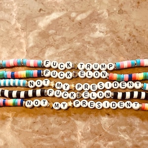 May include: Multicolored beaded bracelets with the phrases "F*ck Trump", "F*ck Elon", "Not My President" repeated on each bracelet.
