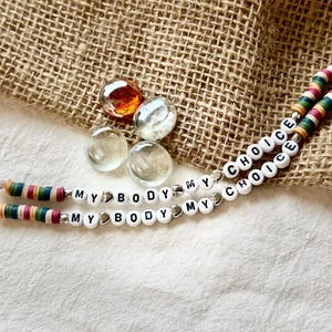 May include: Two beaded necklaces with the words "MY BODY MY CHOICE" and "CHOICE" spelled out in white letter beads. The necklaces have colorful striped beads. Four clear glass marbles and a brown burlap background.