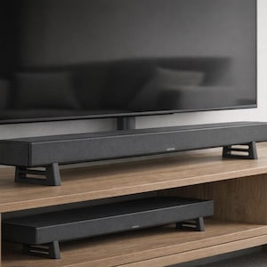 May include: A black soundbar is positioned on a wooden TV stand, beneath a large black television. The soundbar is supported by two black stands. A smaller soundbar is on the shelf below. A small potted plant and books are on the stand.
