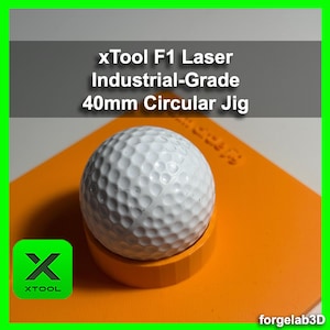 Op de afbeelding: Een witte golfbal ligt op een oranje ronde jig met de tekst "xTool F1 Laser Industrial-Grade 40mm Circular Jig" erboven. De jig is gemaakt door xTool.