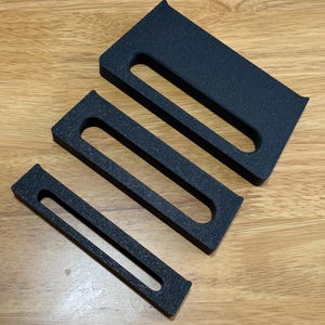 May include: Three black foam inserts of different sizes with a rectangular shape and a cutout in the middle. The inserts are designed to fit inside a box or container.