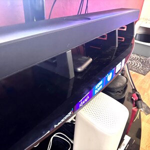 Pu&ograve; includere: Una lunga soundbar grigio scuro &egrave; appoggiata su una superficie nera e lucida. La soundbar &egrave; rettangolare e sembra essere in metallo. La superficie sottostante ha un display digitale con testo colorato. &Egrave; visibile una scatola bianca con piccoli fori.