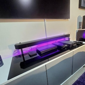 May include: A contemporary entertainment unit featuring a black soundbar and several electronic devices. The setup includes a TV, a media player, and other components, all arranged on a black and grey cabinet with purple lighting.