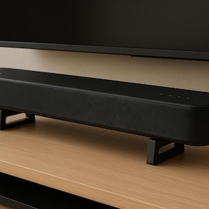 Peut inclure: Une barre de son noire de forme rectangulaire, posée sur deux supports noirs. La barre de son est placée sur une surface en bois brun clair, avec une télévision noire en arrière-plan.