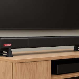 Pu&ograve; includere: Una soundbar nera con la scritta "LG 806AR" su un'etichetta rossa, appoggiata su due supporti neri. La soundbar &egrave; posizionata davanti a un televisore nero e si trova su un mobile in legno.