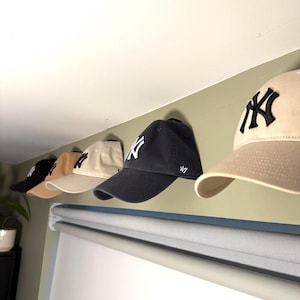 Puede incluir: Cinco gorras de béisbol colgadas de ganchos en una pared. Las gorras son de varios colores, incluyendo negro, beige y azul marino. Una gorra tiene el texto "47" en el lateral.