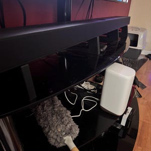 Pu&ograve; includere: Una soundbar nera &egrave; posizionata sopra un mobile porta TV nero, sotto una grande televisione che mostra i loghi dei servizi di streaming. Sono visibili anche un router bianco e uno spolverino grigio. Lo spolverino ha un manico in legno con la scritta "MR.SIGA".