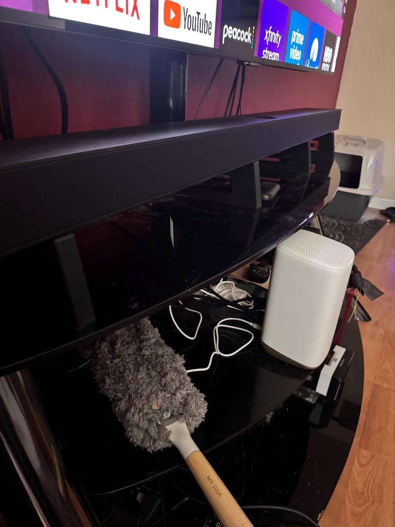 Pu&ograve; includere: Una soundbar nera &egrave; posizionata sopra un mobile porta TV nero, sotto una grande televisione che mostra i loghi dei servizi di streaming. Sono visibili anche un router bianco e uno spolverino grigio. Lo spolverino ha un manico in legno con la scritta "MR.SIGA".