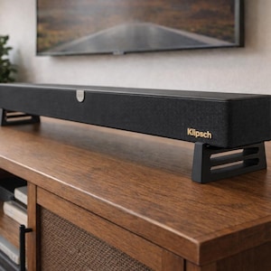 May include: A black soundbar with the brand name "Klipsch" in gold lettering, resting on two black stands. The soundbar is rectangular and sits on a wooden cabinet. A television is visible in the background.