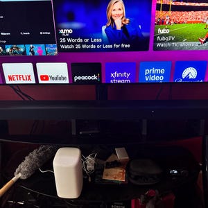 Pu&ograve; includere: Una soundbar nera &egrave; posizionata sotto un grande televisore che mostra i loghi dei servizi di streaming. La TV &egrave; montata sopra una mensola di vetro nero, che contiene un altoparlante bianco, uno spolverino e altri dispositivi elettronici.