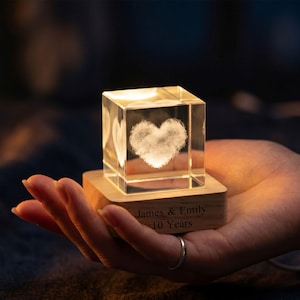 May include: A lit crystal cube with a heart design, set on a wooden base. The base is engraved with the names "James & Emily" and "10 Years". The cube is illuminated from within, creating a warm glow. The cube is likely made of glass.