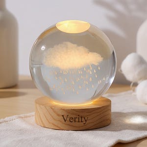 Peut inclure: Une sphère en verre transparent avec un motif de nuage et de pluie, éclairée de l'intérieur, repose sur une base en bois. La base porte l'inscription "Verity". L'objet est décoratif.
