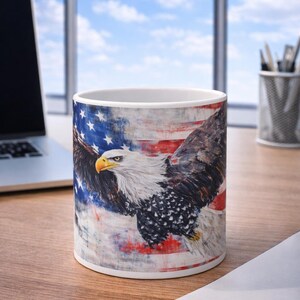 May include: White ceramic mug featuring a majestic bald eagle with outstretched wings, set against a backdrop of the American flag. The design incorporates red, white, and blue colors, creating a patriotic theme. The mug is on a wooden surface.