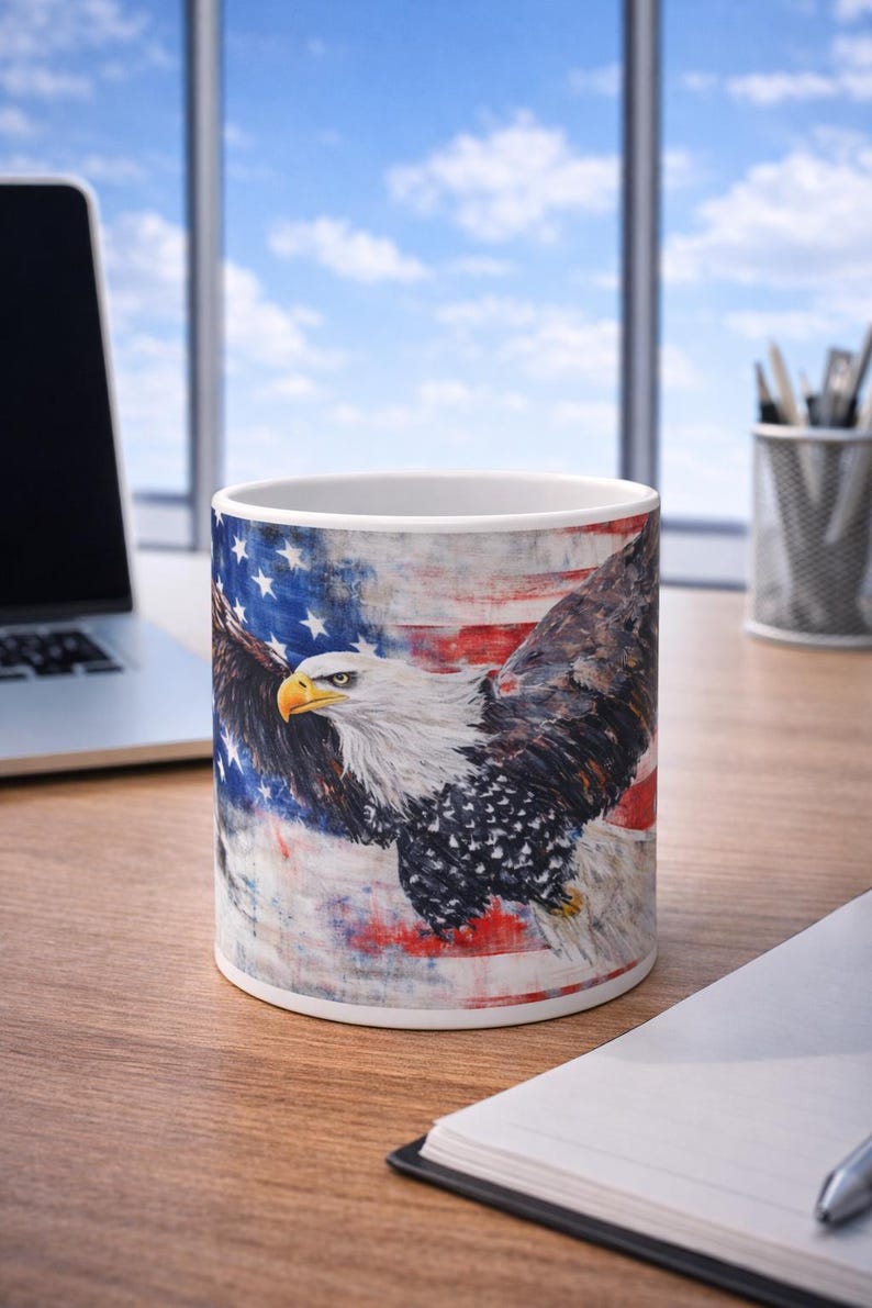 May include: White ceramic mug featuring a majestic bald eagle with outstretched wings, set against a backdrop of the American flag. The design incorporates red, white, and blue colors, creating a patriotic theme. The mug is on a wooden surface.