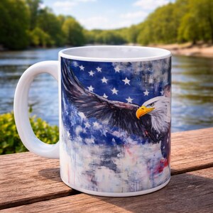 May include: White ceramic mug featuring a majestic bald eagle with outstretched wings against a backdrop of the American flag. The mug has a white handle and is set on a wooden surface, perfect for patriotic displays.