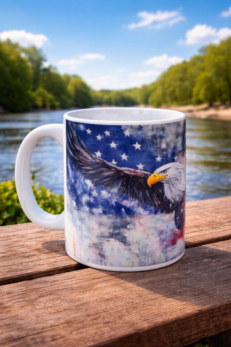 May include: White ceramic mug featuring a majestic bald eagle with outstretched wings against a backdrop of the American flag. The mug has a white handle and is set on a wooden surface, perfect for patriotic displays.