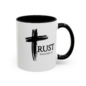 May include: A white ceramic mug with a black interior and handle. The exterior features a black brushstroke cross graphic with the word TRUST in black serif font, followed by the text Proverbs 3:5 in a smaller black serif font below it.