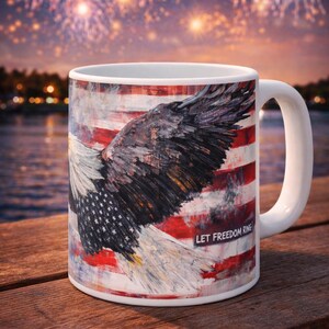 May include: White ceramic mug featuring an American flag design with a bald eagle and the text "LET FREEDOM RING." The mug has a white handle and is set against a blurred background of fireworks.