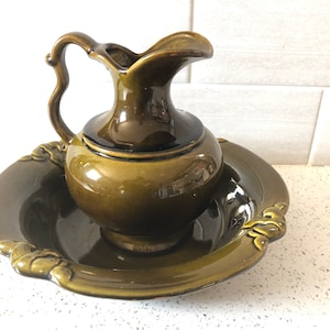 Puede incluir: Juego de jarra y palangana de cerámica verde oliva. La jarra tiene un asa curva y un pico. La palangana redonda presenta detalles decorativos. El conjunto es probablemente vintage y adecuado para uso decorativo.