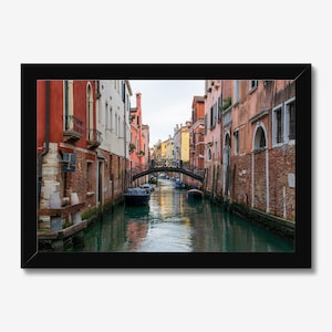 May include: A framed photograph of a Venetian canal scene. The image features a narrow waterway lined with colorful buildings in shades of red, orange, and white. A decorative bridge arches over the water, and several boats are docked along the canal.