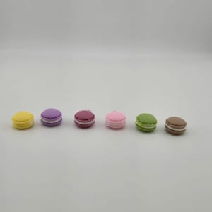 Könnte beinhalten: Reihe von sechs bunten Macarons in Gelb, Lila, Burgunderrot, Rosa, Grün und Braun. Jedes Macaron hat eine weiße Füllung und eine glatte, runde Form, was auf ein Dessert-Thema hindeutet.