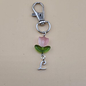 Puede incluir: Un llavero plateado con un charm de tulipán de vidrio rosa y verde y un charm de letra L plateado.