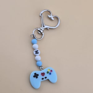 Puede incluir: Un llavero de plástico azul en forma de mando de videojuego con un corazón y botones coloridos. El llavero tiene un cierre de corazón plateado y una cadena de cuentas blancas y azules que deletrean la letra "M".