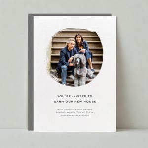 May include: A housewarming invitation card with a photo of a couple and their dog on a set of stairs. The text reads "You're invited to warm our new house with laughter and drinks Sunday, March 7th at 5 p.m. Our brand new place."