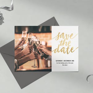 Op de afbeelding: Een save-the-date kaart met gouden belettering en een foto van mensen die proosten met champagneglazen. De kaart ligt op een grijze envelop, met een zilveren strik en lint.