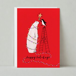 May include: A Christmas card with a red background features a polar bear wrapped in string lights and a penguin on a ladder placing a star on top. The words "happy holidays" are written in cursive.