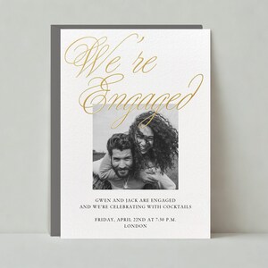 以下が含まれることがあります： 婚約を発表する白いカードに金色の文字が書かれています。テキストには「We're Engaged」とあります。カードの中央には、カップルが写った白黒写真があります。写真のすぐ下には、「Gwen and Jack are engaged and we're celebrating with cocktails Friday, April 22nd at 7:30 p.m. London.」というテキストが書かれています。
