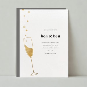 May include: A white invitation card with gold foil accents. The card features a champagne flute with bubbles rising from the top. The text on the card reads "JOIN US AS WE TOAST bea & ben ON THEIR 5TH ANNIVERSARY AS HUSBAND AND WIFE SATURDAY, SEPTEMBER 16TH AT 6PM NORWOOD CLUB".