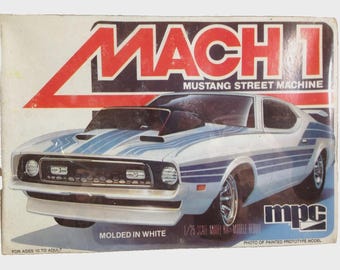 May include: A model kit box for a Mach 1 Mustang Street Machine. The box features a white car with blue stripes and the text "MACH 1" in red. The box also says "MOLDED IN WHITE" and "1/25 SCALE MODEL KIT".
