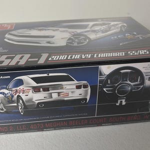 Puede incluir: Caja de modelo de un Chevy Camaro SS/RS Coupe de 2010. La caja presenta im&aacute;genes del coche blanco con gr&aacute;ficos con tem&aacute;tica de la bandera estadounidense. El texto "USA-1" es visible.