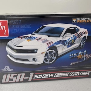 Puede incluir: Caja de modelo de un Chevrolet Camaro SS/RS Coupe blanco de 2010 con gr&aacute;ficos patri&oacute;ticos de EE. UU. El coche tiene detalles en azul y rojo con el texto "USA-1". La caja incluye el texto "1/25" y "Kit de pl&aacute;stico".