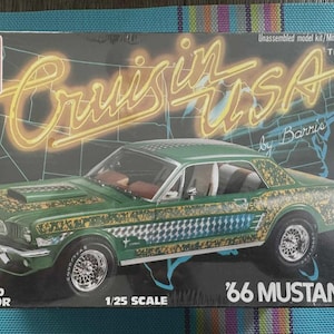 Puede incluir: Caja de modelo de un coche Mustang '66 verde con detalles dorados y a cuadros. La caja tiene el texto "Cruisin' USA" en una fuente de estilo ne&oacute;n, y "1/25 Scale" est&aacute; impreso en la caja.
