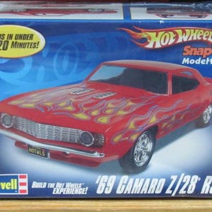May include: A red '69 Camaro Z/28 RS model kit with yellow flame decals. The box features the Hot Wheels logo and text that reads "Builds in under 20 minutes!" and "Snap Tite Model Kit". The Revell logo is also visible.