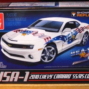 Puede incluir: Maqueta de un Chevy Camaro SS/RS Coupe 2010 blanco, decorado con gr&aacute;ficos patri&oacute;ticos con tem&aacute;tica de EE. UU. La caja presenta el coche, una estrella y la bandera estadounidense. El modelo es a escala 1/25.