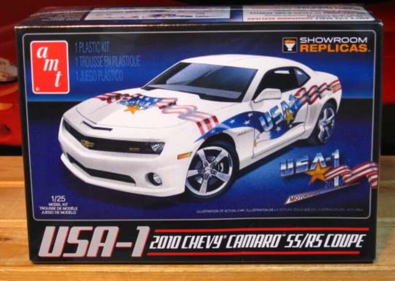 Puede incluir: Maqueta de un Chevy Camaro SS/RS Coupe 2010 blanco, decorado con gr&aacute;ficos patri&oacute;ticos con tem&aacute;tica de EE. UU. La caja presenta el coche, una estrella y la bandera estadounidense. El modelo es a escala 1/25.