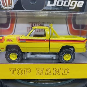 May include: Yellow Dodge pickup truck toy with black tires and a roll bar. The truck has orange and red stripes and the text "TOP HAND" on the side. The toy is in a clear plastic display case.