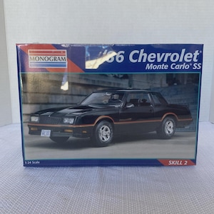 May include: A model kit box for a black '86 Chevrolet Monte Carlo SS. The box features a picture of the car with orange trim, the Monogram logo, and the text "1:24 Scale" and "Skill 2".