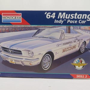 May include: A model kit box featuring a white '64 Mustang Indy Pace Car. The box has a blue background with the Monogram logo and the text "'64 Mustang Indy Pace Car." The car model is a convertible.