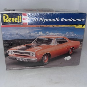 May include: A Revell 70 Plymouth Roadrunner plastic model kit in a rectangular box. The box features an orange car with a black hood stripe, 1:24 scale, and skill level 2. The text reads Revell, 70 Plymouth Roadrunner, 1:24 Scale, Ages 10 & Up, Skill Level 2.